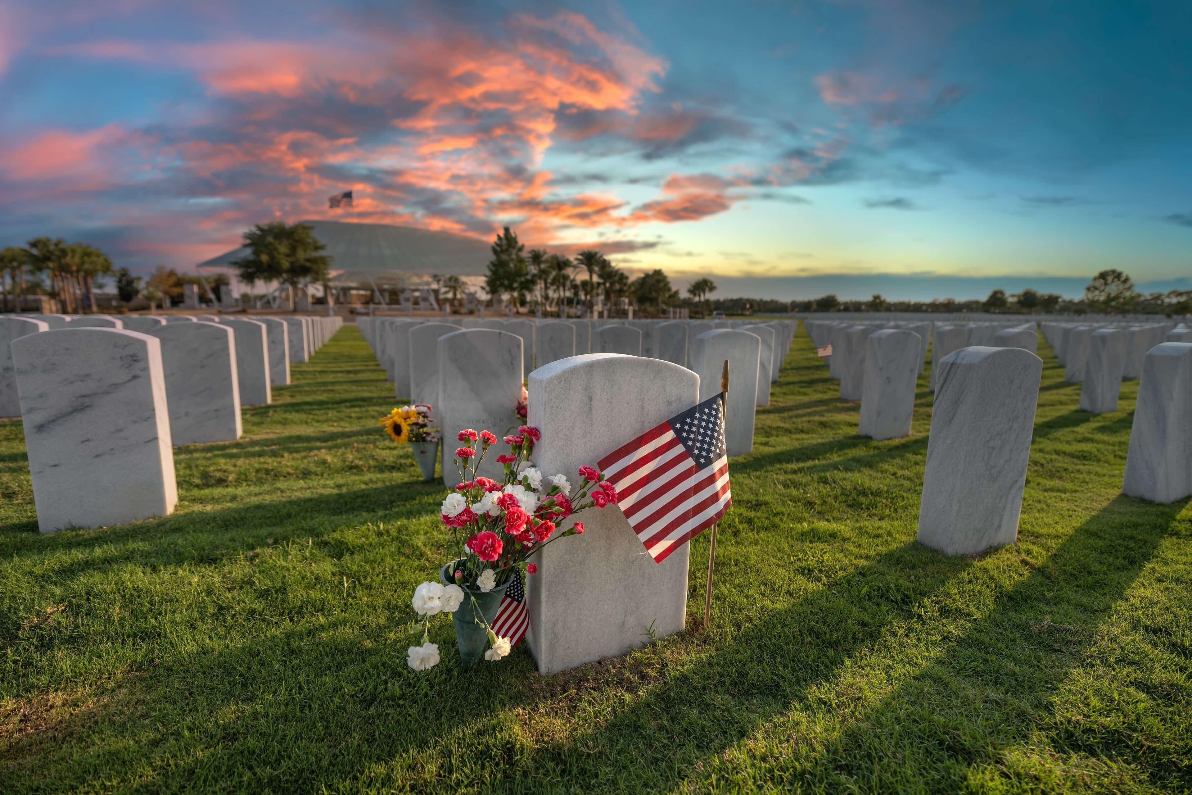A grave adorned with flowers and an American flag, surrounded by gravestones under a colorful sunset. A grave adorned with flowers and an American flag, surrounded by gravestones under a colorful sunset.