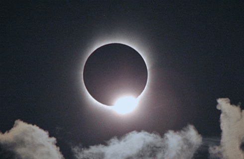 Moon covering the sun with a small white ring around it and clouds below.
