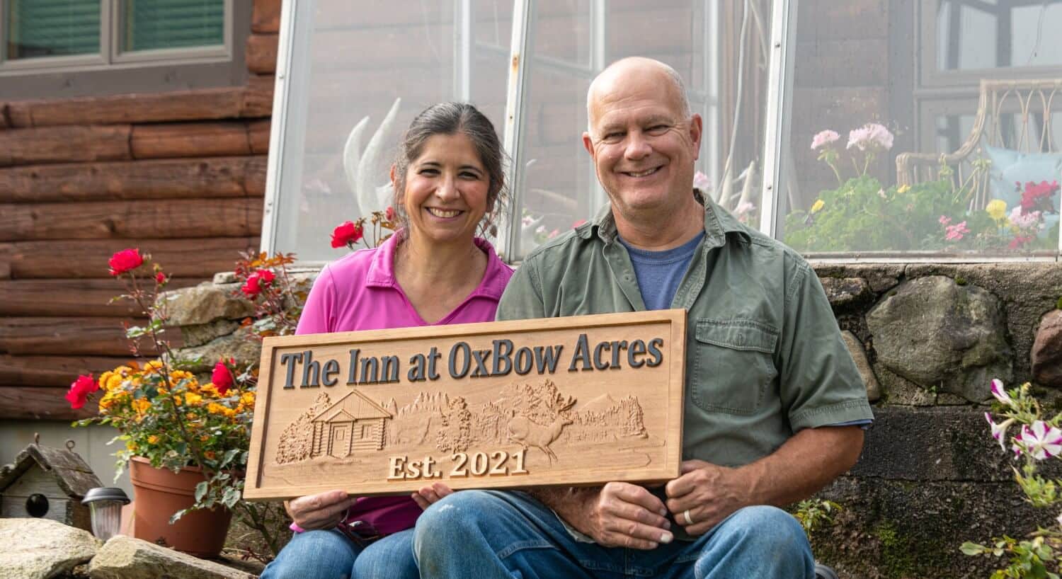 Woman in pink shirt and man in green shirt holding a wooden sign that says The Inn at Oxbow Acres.