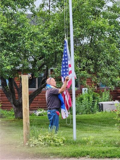 A man in with sunglasses on his head, raising the flag on a pole next to a hitching post.