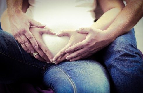 Pregnant woman holding her hands in the shape of a heart on her belly with a man's arms around her
